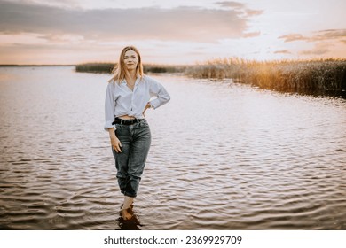 A woman in jeans and a white shirt stands in the water at sunset on the shore of a lake - Powered by Shutterstock