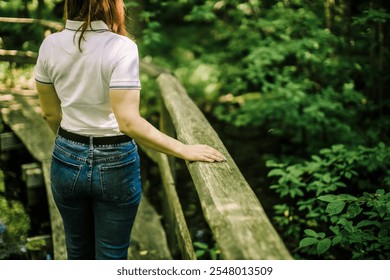 A woman in jeans and a white shirt touches a rustic wooden bridge railing, surrounded by lush forest greenery. A serene and tranquil outdoor moment ideal for nature and lifestyle themes - Powered by Shutterstock