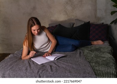 Woman In Jeans And A White Top Lying On Her Side In Bed And Writing Notes In A Notebook