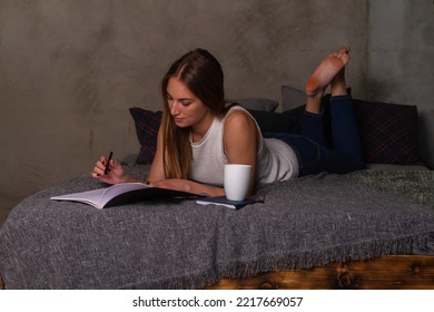 Woman In Jeans And A White Top Lying On Her Stomach In Bed And Writing Notes In A Notebook