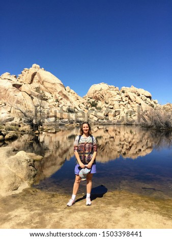 A woman at Joshua Tree National Park standing in front of rock formations reflecting in a lake and wearing a sweater of buildings reflecting in water. She is holding a hat and wearing a backpack.
