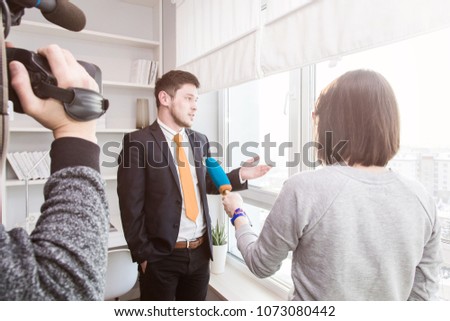 A woman journalist interviews a business man in a suit near a window in a room with a modern interior