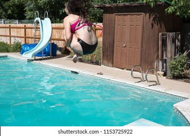 Woman Jumping Off Diving Board Doing A Cannon Ball Into A Private Swimming Pool. Young Woman Mid Air Doing A Cannon Ball Into An Outdoor Swimming Pool In The Summer. Summer Fun In The Pool.