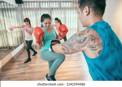 Woman Kick Boxer Kicking Her Trainer In A Sparring Session. In The Background Other Boxers Are Practicing Hitting Punching Pose In Air. Group Of Asian Girls Taking Boxing Lesson With Coach In Gym.