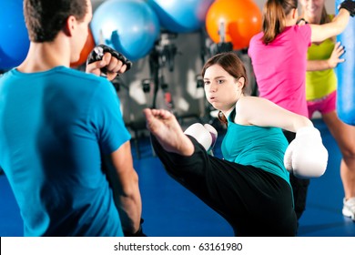 Woman Kick Boxer Kicking Her Trainer In A Sparring Session, In The Background Other Boxers Are Hitting The Sandbag