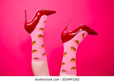 A Woman Kicks Her Feet Up In The Air, She Is Wearing Red High Heel Stilettos And Funny Pink Socks With Burgers Printed On Them. 