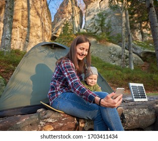 Woman And Kid Take A Selfie On A Mobile Phone On The Background Of A Tourist Tent With A Solar Panel At The Foot Of A Cliff In The Forest. Family In The Sunbeams With Gentle Smiles