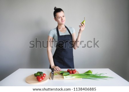 A woman in a kitchen apron holds a green pear on a gray background. On the white table are the vegetables.