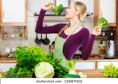 Woman In Kitchen With Many Green Leafy Vegetables, Fresh Produce On Counter. Young Housewife Having Fun Holds Broccoli., Singing. Healthy Eating, Cooking, Vegetarian Food, Dieting Concept.