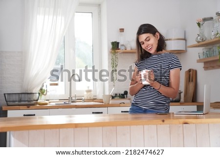A woman in the kitchen with a smile. A woman drinks coffee and holds a cup in her hand. She is happy because She drinks her favourite coffee in the morning. High quality photo with copy space.