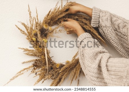 Woman in knitted sweater holding autumn wreath and decorating house entrance. Stylish rustic autumn wreath with dried grass and berries for farmhouse front door