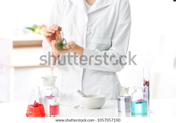 Woman in lab coat holding glass bottle with perfume oil near table indoors