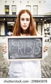 Woman in a lab coat on a cobblestone street. with a serious look on her face holding a chalkboard with the word "CRISIS" on it. Processed for dramatic effect. - Shutterstock ID 298845878