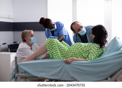 Woman With Labor Contractions Giving Birth To Baby In Hospital Ward At Maternity Clinic. Obstetrics Doctor And African American Nurse Assisting Child Delivery With Young Husband