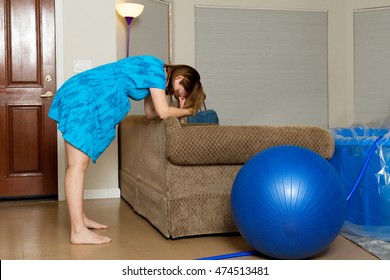 A woman in labor leans forward onto the back of a couch as she breathes through a contraction.  There is a birthing ball and a birthing tub in the room.