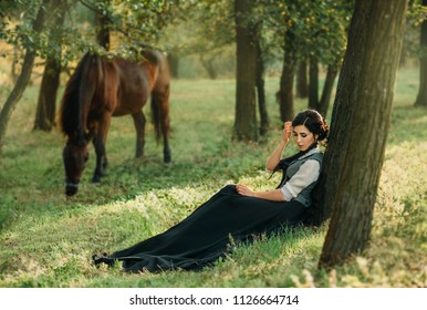 Woman lady princess. vintage black dress costume medieval clothes sits rests dreaming enjoy forest summer nature under tree sunny day. background walks brown animal horse. Fantasy photography quuen 