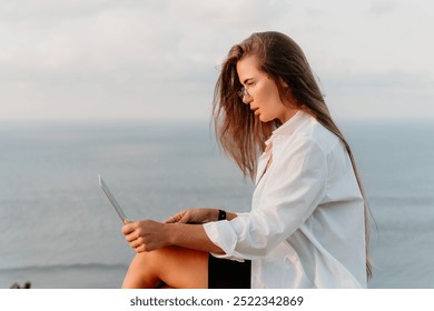 Woman Laptop Cliffside Ocean - A woman works on her laptop while sitting on a cliffside overlooking the ocean. - Powered by Shutterstock