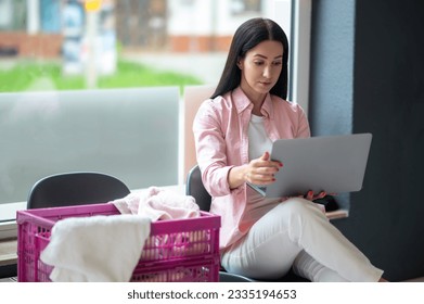 Woman with laptop at laundromat doing job project, connection and multitask, working on computer while washing clothes. - Powered by Shutterstock