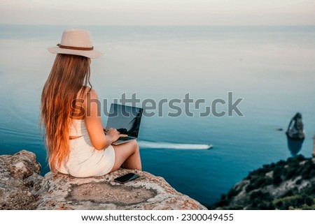 Woman laptop sea. Working remotely on seashore. Happy successful woman female freelancer in straw hat working on laptop by the sea at sunset. Freelance, remote work on vacation