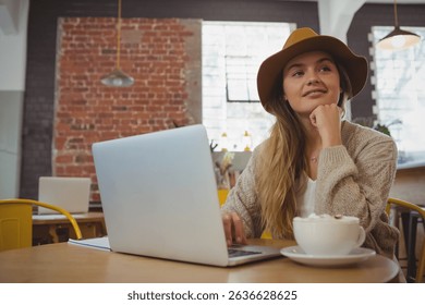 Woman in late twenties working on silver laptop at café table with coffee cup and notebook. Urban, hospitality, modern, workspace, cozy, creative, lifestyle