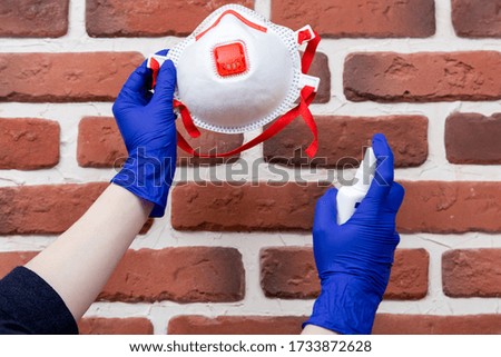 Woman in latex gloves is cleaning her disposal respirator using sanitizer. Reusing of respirators in the context of their deficit and high cost. Brick wall background.