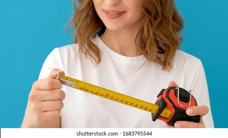 Woman Laughs At A Small Size And Enlarges It To A Larger. Female In White T Shirt Unrolling Construction Measuring Tape Against Blue Background