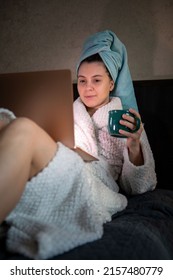 Woman Laying Down In Bed With Wet Head Covered With Towel Working On Laptop Drinking Tea