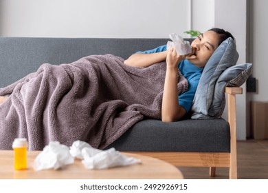 A woman is laying on a couch with a tissue in her mouth. She is wearing a blue shirt and has a tissue on her face. The couch is covered in a blanket and there is a bottle of medicine nearby - Powered by Shutterstock