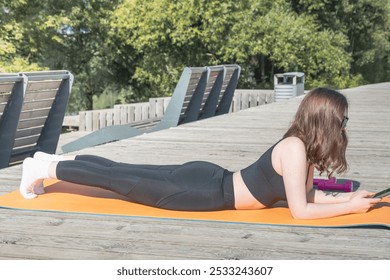 A woman is laying on a yoga mat on a wooden deck. She is reading a book and wearing glasses - Powered by Shutterstock