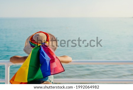 a woman leaning on a railing looks out to sea with a rainbow-colored scarf. lgtb. pride flag.