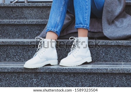 Woman legs in blue jeans and white shoes with laces. Stylish woman sitting on the stairs. Street fashion look, winter outfit.
