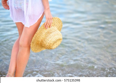 Woman Legs And A Straw Hat In Hand On The Beach In Sea Water. Summer Vacations Concept