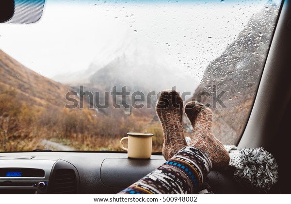 Woman legs in warm socks on car\
dashboard. Drinking warm tee on the way. Fall trip. Rain drops on\
windshield. Freedom travel concept. Autumn weekend in\
mountains.