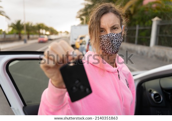 Woman with leopard skin face mask holding card\
key by white vehicle with open door. Car rental, free ride,\
insurance concepts