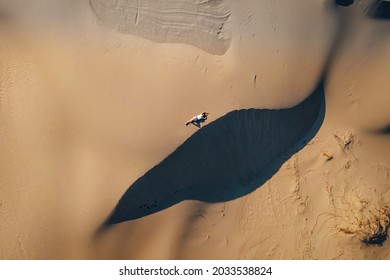 Woman Lie On Wavy Sand Dunes In Desert Landscape At Golden Sunset Light And Wind.