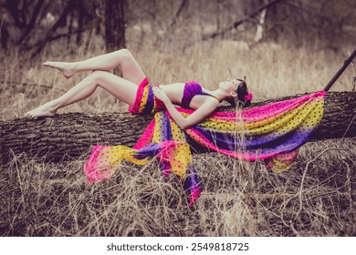 A woman lies gracefully on a fallen tree in a meadow, draped in vibrant purple, pink, and yellow fabric, exuding serenity and whimsy. Perfect for artistic and bohemian themes - Powered by Shutterstock