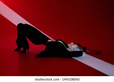 Woman Lies On Red Background With Ray Of Light On Face. Sexy Nude And Confident Blonde Woman In Black Jacket. Red Light
