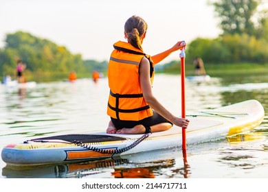 woman in life jacket sit at sub board at river ar evening , forest trees background