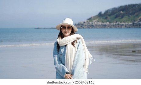 A woman in a light blue coat, white scarf and hat, stands on a beach with calm sea, rocky shoreline, and a hilly backdrop. - Powered by Shutterstock