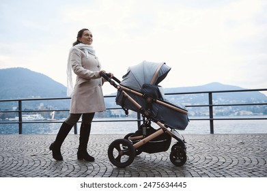 A woman in a light coat and scarf walks with a baby stroller along a lakeside path. Serene landscape with mountains in the background, suggesting peacefulness and care. - Powered by Shutterstock