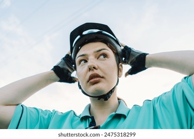 A woman in a light green shirt and black gloves adjusts her black bike helmet while looking upwards. The image captures a moment of preparation before heading out on a bike ride. - Powered by Shutterstock