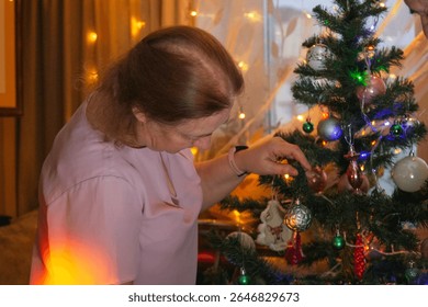 A woman in a light purple shirt carefully hangs colorful ornaments on a Christmas tree in a warmly lit living room