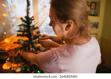 A woman in a light purple shirt carefully hangs colorful ornaments on a Christmas tree in a warmly lit living room