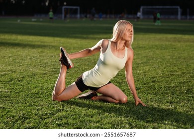 A woman in a light tank top performs a quad stretch on a grassy field, showcasing flexibility and focus during an outdoor fitness session, with a blurred sports background - Powered by Shutterstock