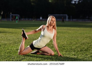 A woman in a light tank top performs a quad stretch on a grassy field, showcasing flexibility and focus during an outdoor fitness session, with a blurred sports background - Powered by Shutterstock