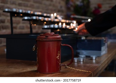 Woman Lights Candle In Church After Throwing Money Into Red Metal Collection Box For Donations. Focus On The Collection Box On The Table