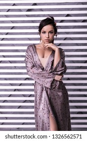 Woman In Lilac Wrap Dress. Charming Radiantly Beautiful Female With Tied Up Brown Hair Wearing Silk Pattern Wrap Dress Posturing Over Striped Background