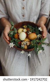 Woman in linen dress holding easter eggs and spring flowers in wooden bowl. Stylish easter and quail eggs in natural dye and spring blooms. Aesthetic holiday. Happy Easter!
