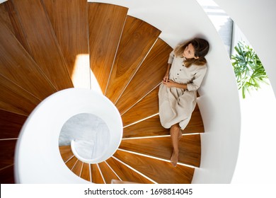 Woman In Linen Natural Dress Is Sitting On The Wooden Modern Stairs Inside Of The House