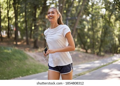 Woman listen music running on asphalt track summer park Caucasian female earphones jogging sunny morning Runner training hold smartphone in hand Healthy lifestyle positive concept  - Powered by Shutterstock